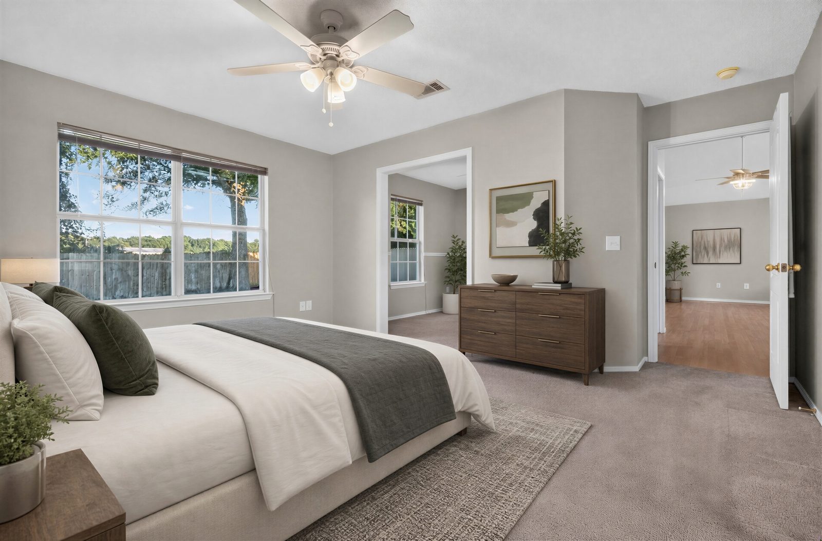 Primary bedroom, virtually staged: a wide window onto the fenced yard and a shade tree, a ceiling fan, and doorways to the office and the hall, shown with digitally added furniture